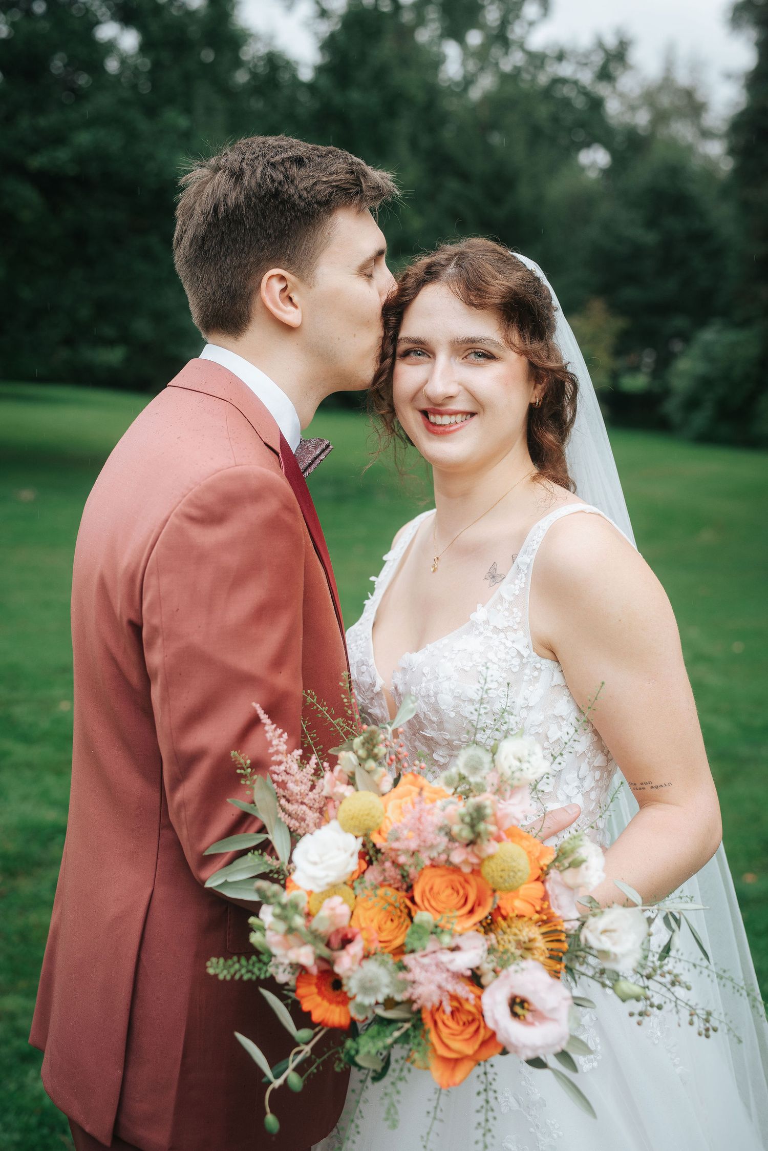 Groom in a rust-colored suit kisses his smiling bride on the forehead as she holds an orange and pink bouquet on a green lawn.