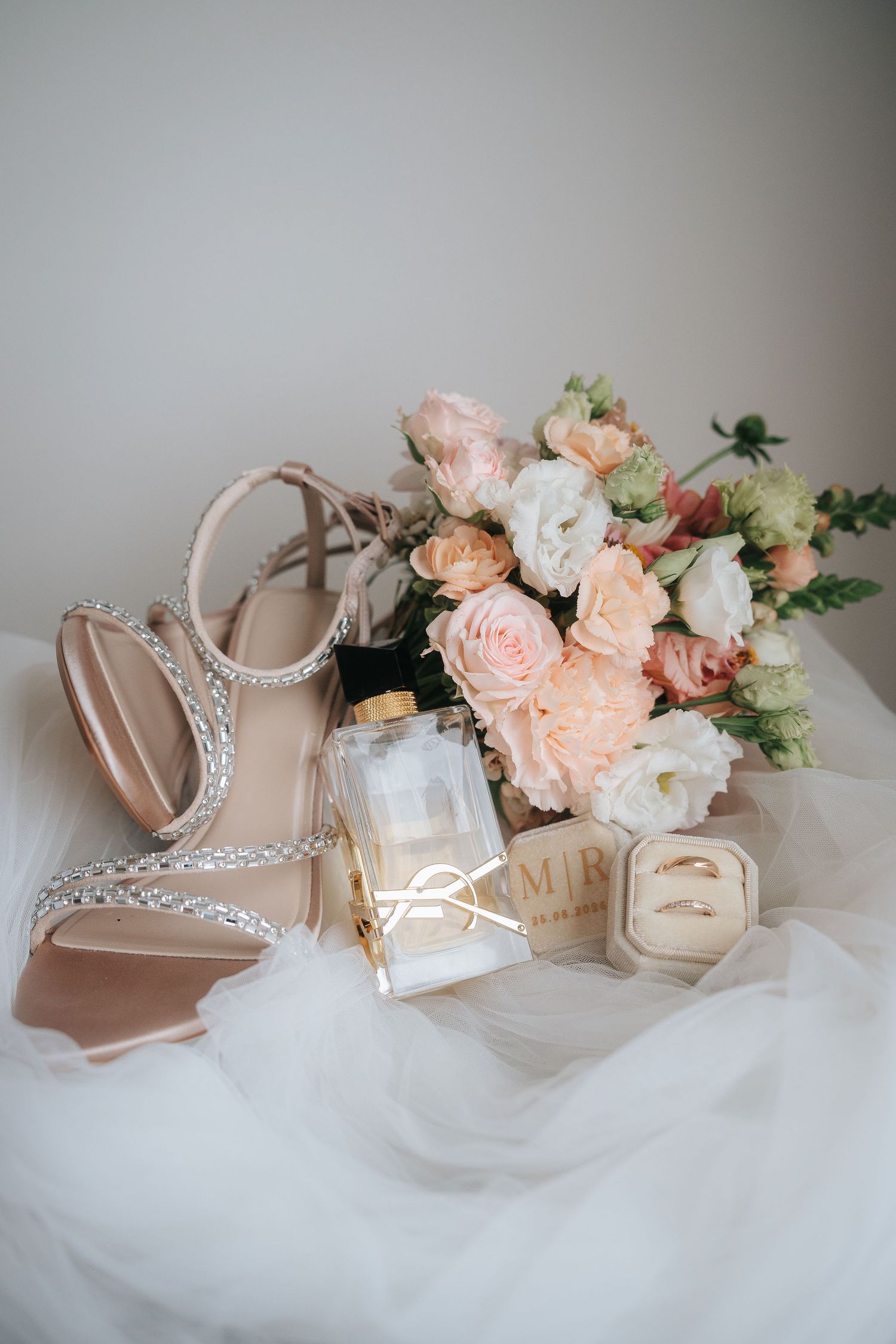 Wedding flat lay with blush rhinestone sandals, perfume bottle, ring box and a peach and white rose bouquet on soft tulle.