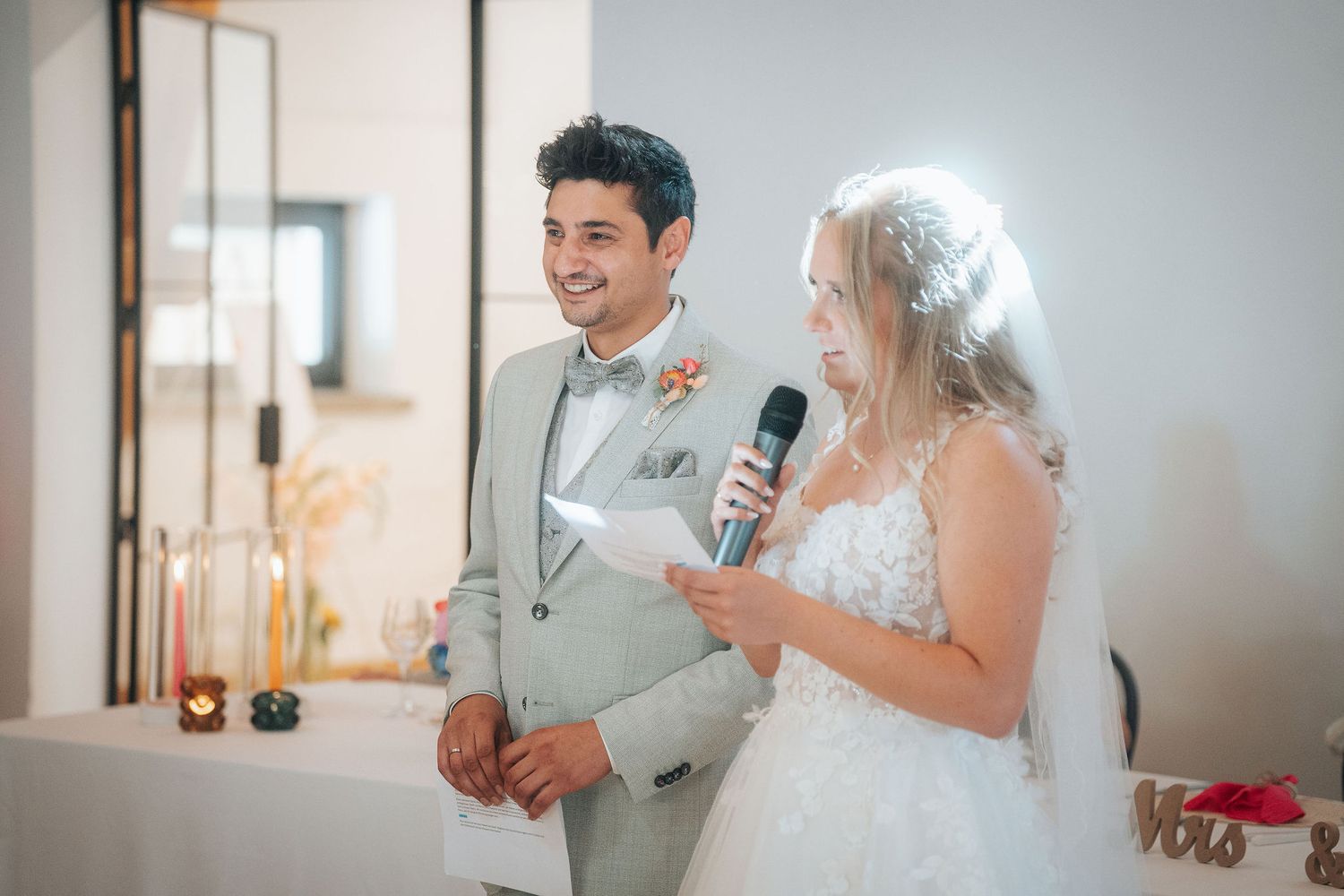 Smiling groom in a gray suit watches as the bride in a lace gown and veil speaks into a microphone at their wedding reception.