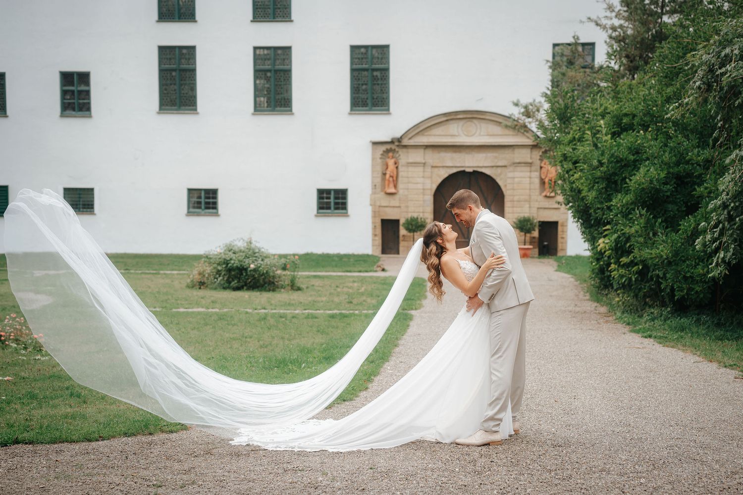 Bride and groom share an intimate embrace on a gravel path with her long veil flowing behind, in front of a historic white building.