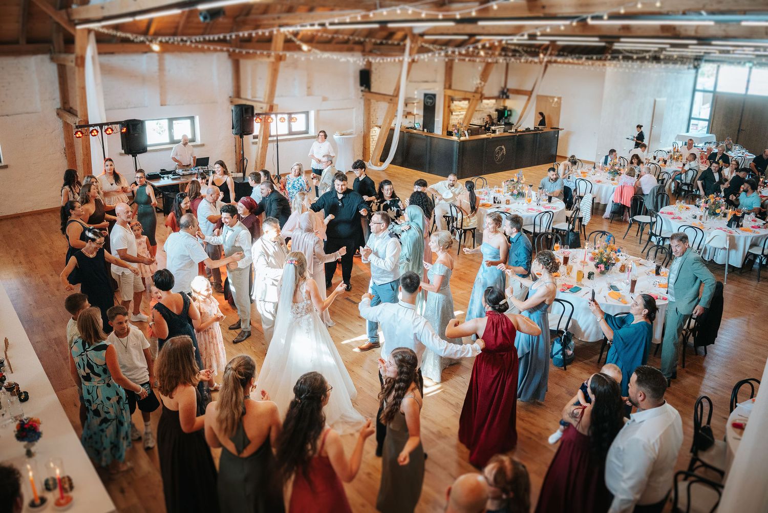 Wedding guests gather around a bride in a white gown on the dance floor of a rustic reception hall with wood beams and string lights.