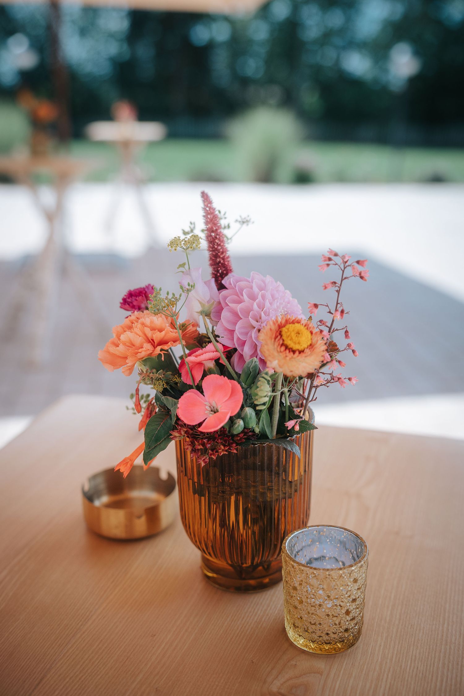 Colorful wedding table centerpiece of pink, coral and orange flowers in an amber ribbed glass vase beside a gold ashtray and candle holder.
