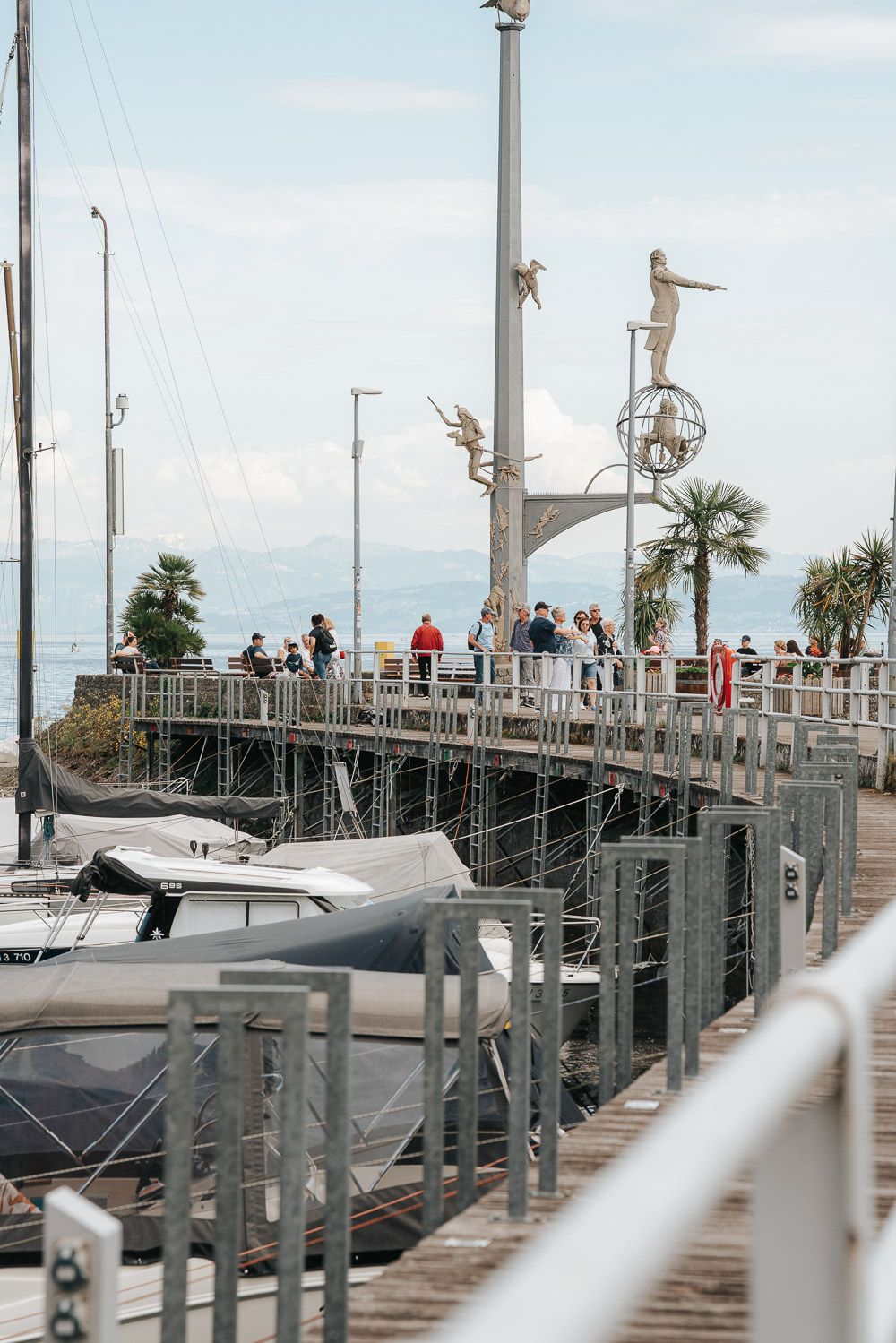 Visitors stroll along a marina pier with boats docked alongside and decorative metal sculptures mounted on light posts.