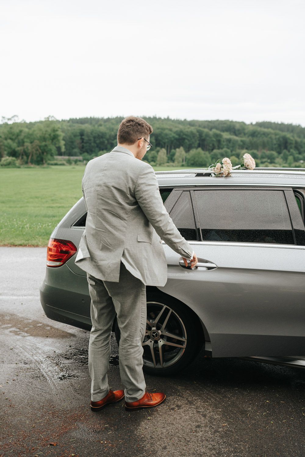 Someone in a light gray suit opens the door of a silver Mercedes station wagon parked on wet ground with green hills behind.