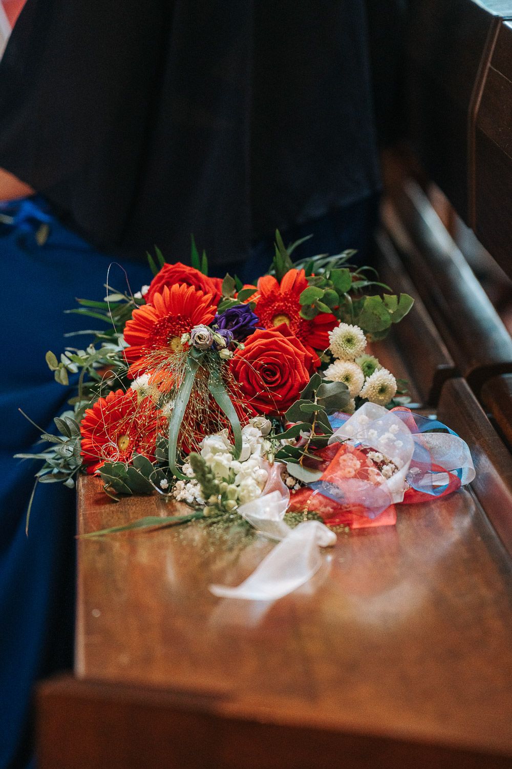 A vibrant floral arrangement with red gerbera daisies and roses rests on a wooden pew or bench.