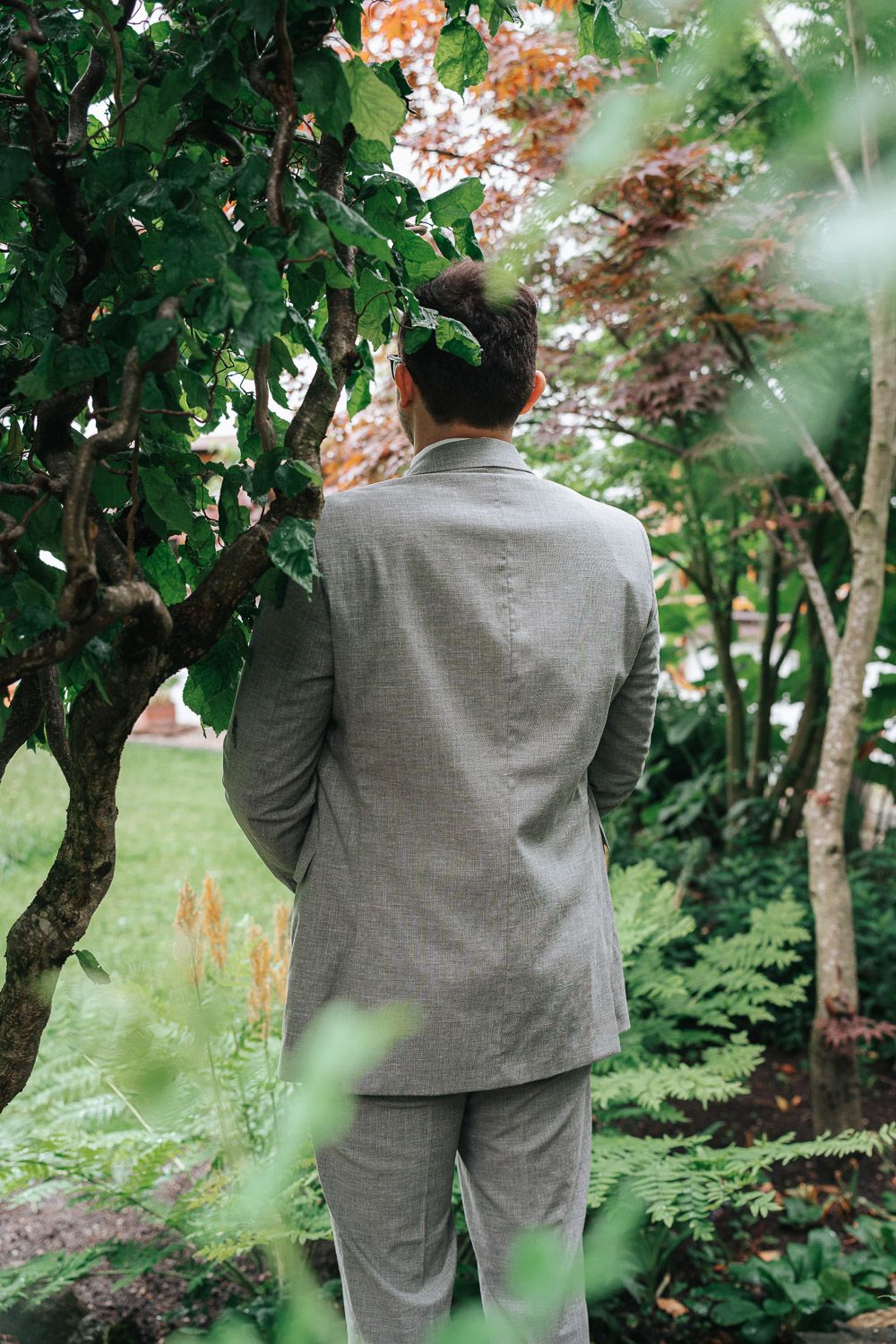 Figure in a light gray suit stands among lush garden greenery and flowering trees on a peaceful summer day.