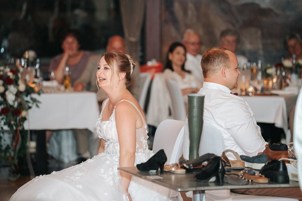 A couple at a wedding reception share a playful moment sitting back-to-back on chairs at an elegant indoor venue.