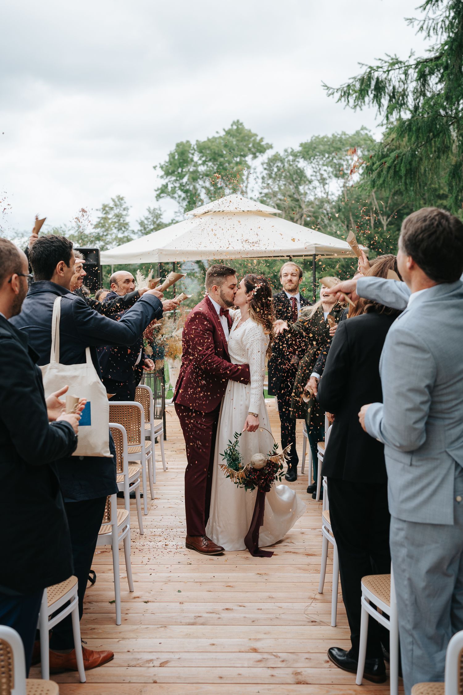 Wedding ceremony guests shower newly married couple with confetti under white gazebo on wooden deck.