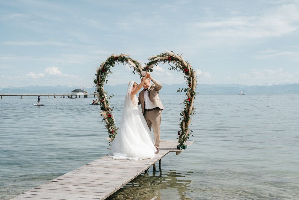 A couple shares a romantic moment under a heart-shaped floral arch on a wooden pier overlooking calm ocean waters.