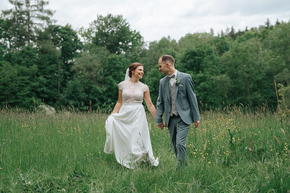 A couple in wedding attire walks together through a lush green meadow on a sunny day.