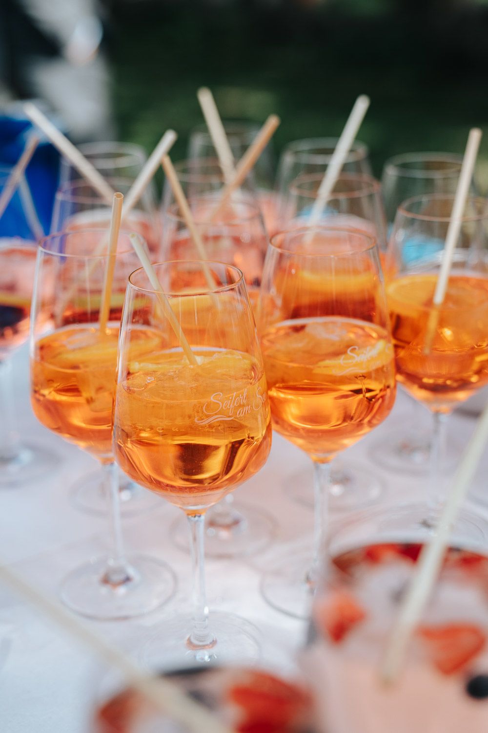 Multiple wine glasses filled with orange-hued cocktails lined up with white straws stand ready for service on a light surface.