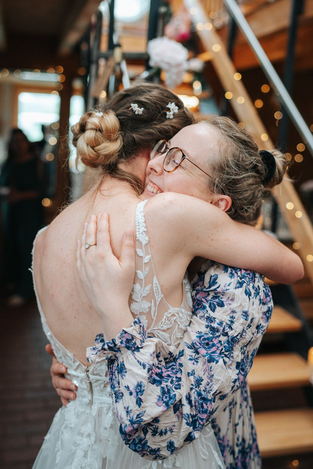 Bride and wedding guest embrace in an emotional hug, with blue floral dress and white lace gown details visible.