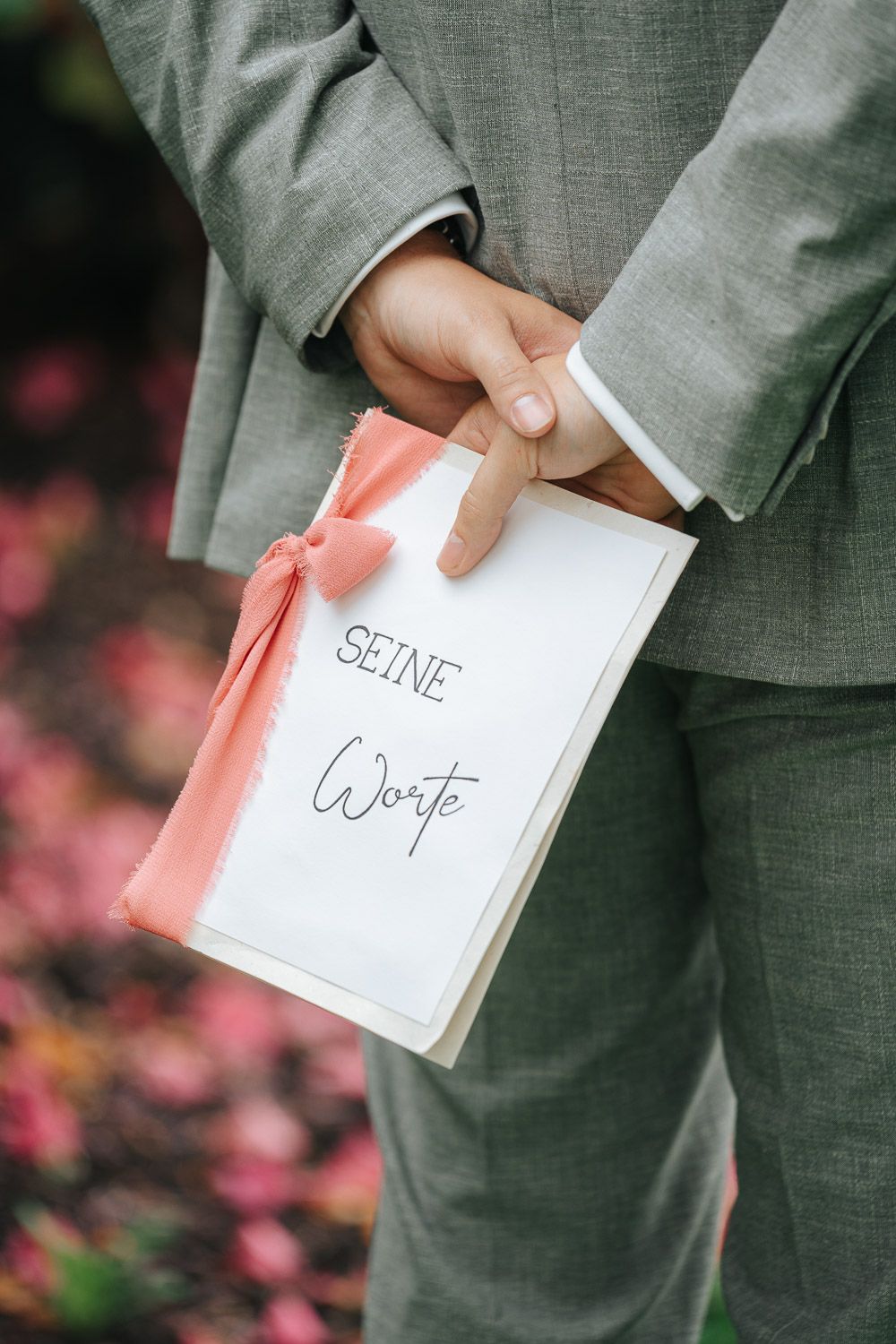 Hands holding white wedding vows booklet with peach ribbon against gray suit and pink floral background.