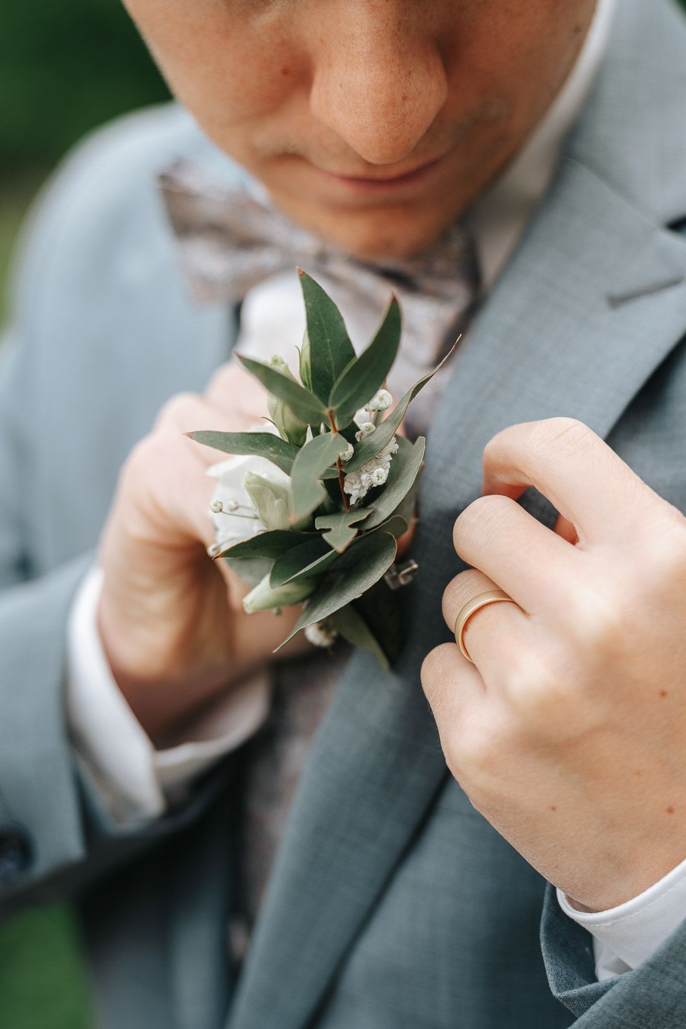 A close-up of hands pinning a delicate white floral and greenery boutonniere onto a gray suit jacket.
