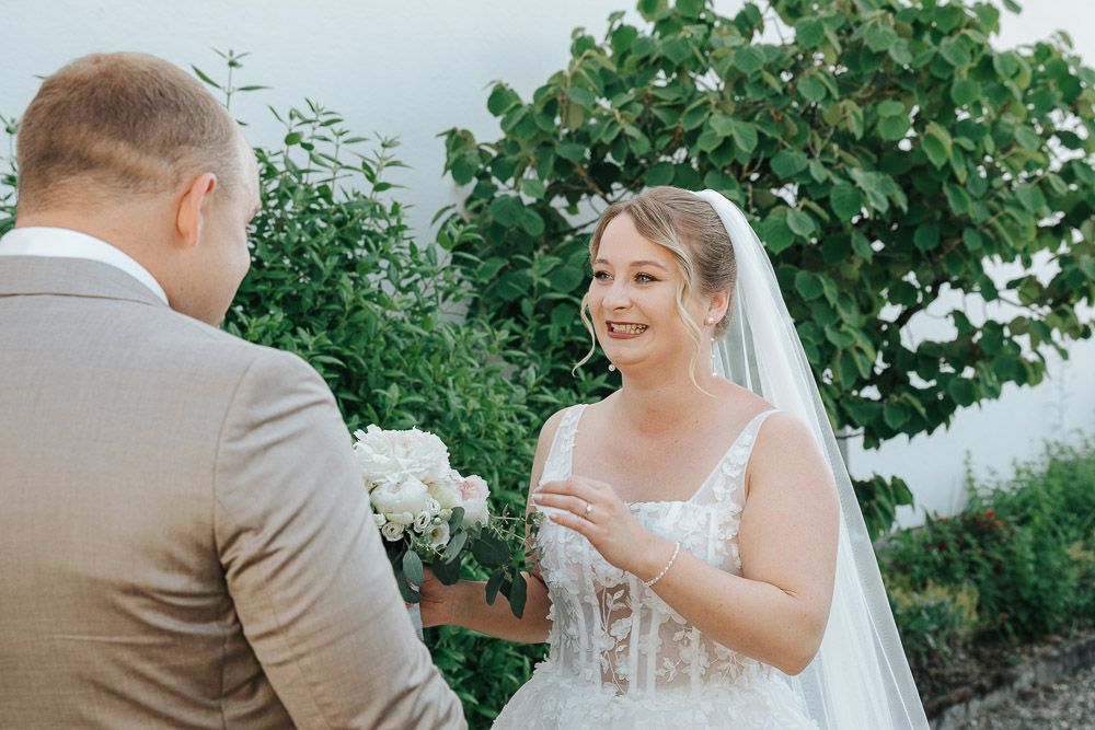 A joyful interaction between a bride in a white lace dress and groom in gray suit during an outdoor wedding ceremony.