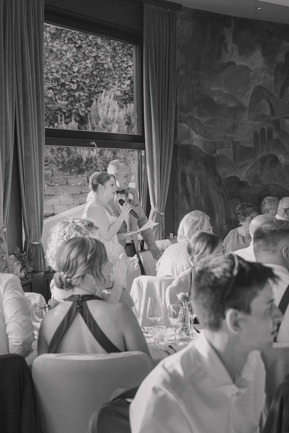 Black and white photo of guests dining in an elegant reception room with curtains and wall murals.