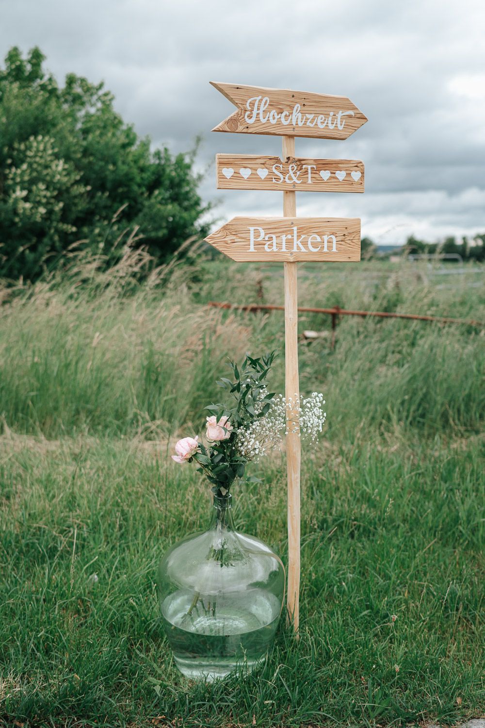 Rustic wooden directional signs for 'Hochzeit' and 'Parken' stand among tall grass with glass vase flower decorations below.