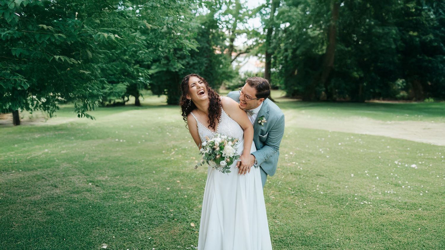A joyful couple shares a candid moment on a green lawn surrounded by trees during their outdoor wedding celebration.