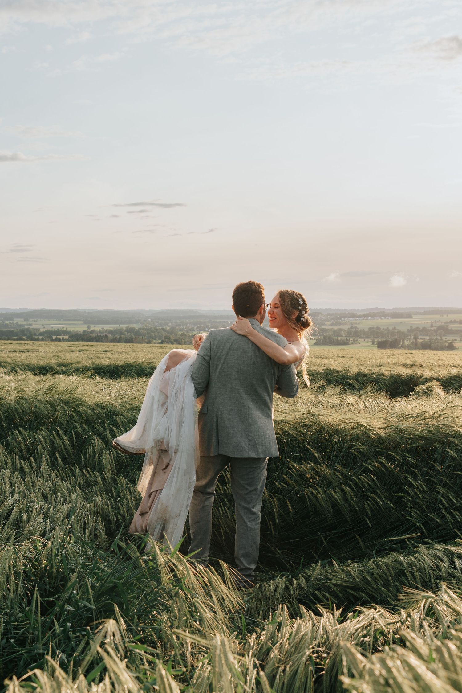 A couple embraces while standing in a golden wheat field at sunset with rolling hills in the background.