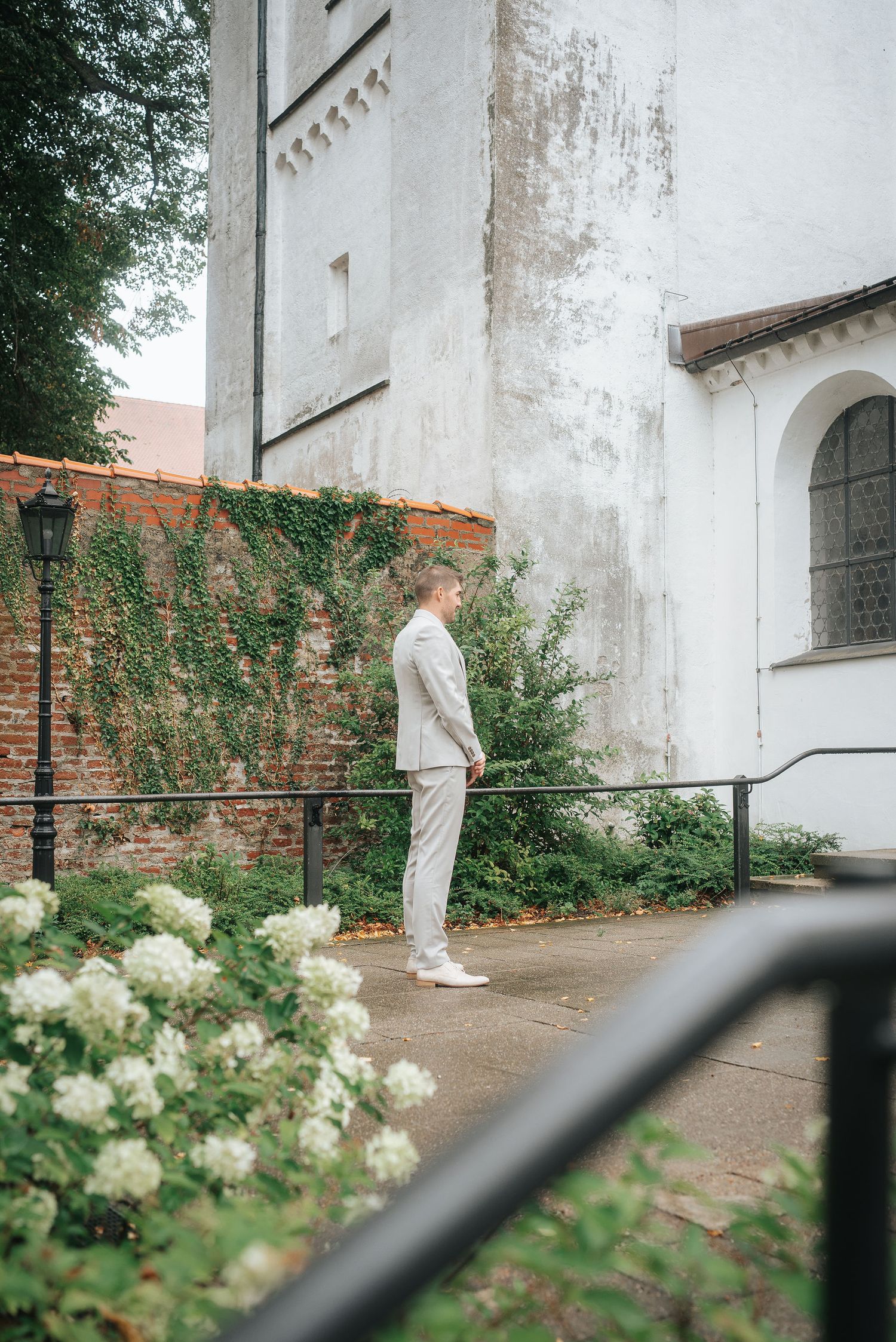 Man in a light gray suit and white shoes stands beside a weathered white church wall, ivy-covered brick, and blooming white hydrangeas.