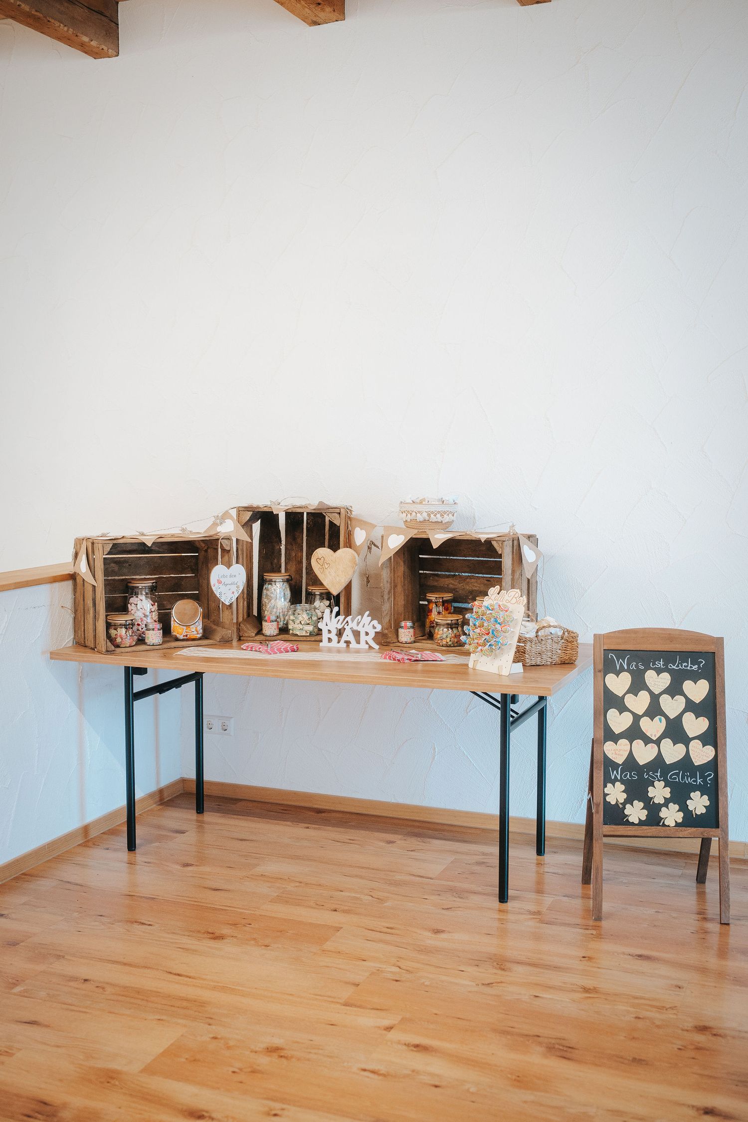 Wooden crates on a rustic table hold a wedding candy bar and bar sign, beside a chalkboard with heart-shaped guest messages.