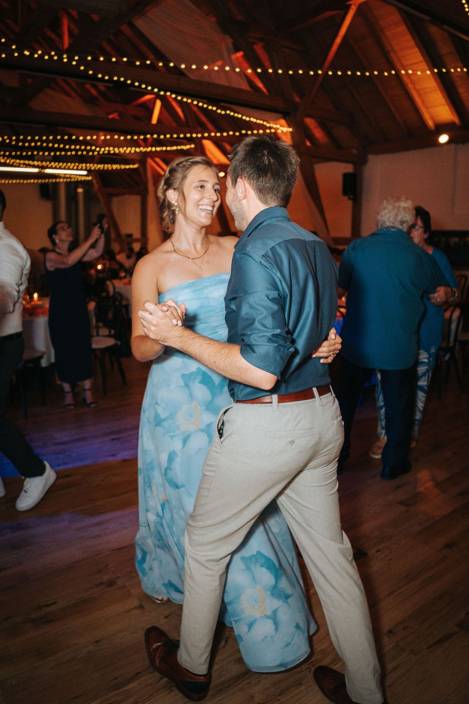 Smiling woman in a light blue floral dress dances with a man in a teal shirt at a lively reception under string lights.