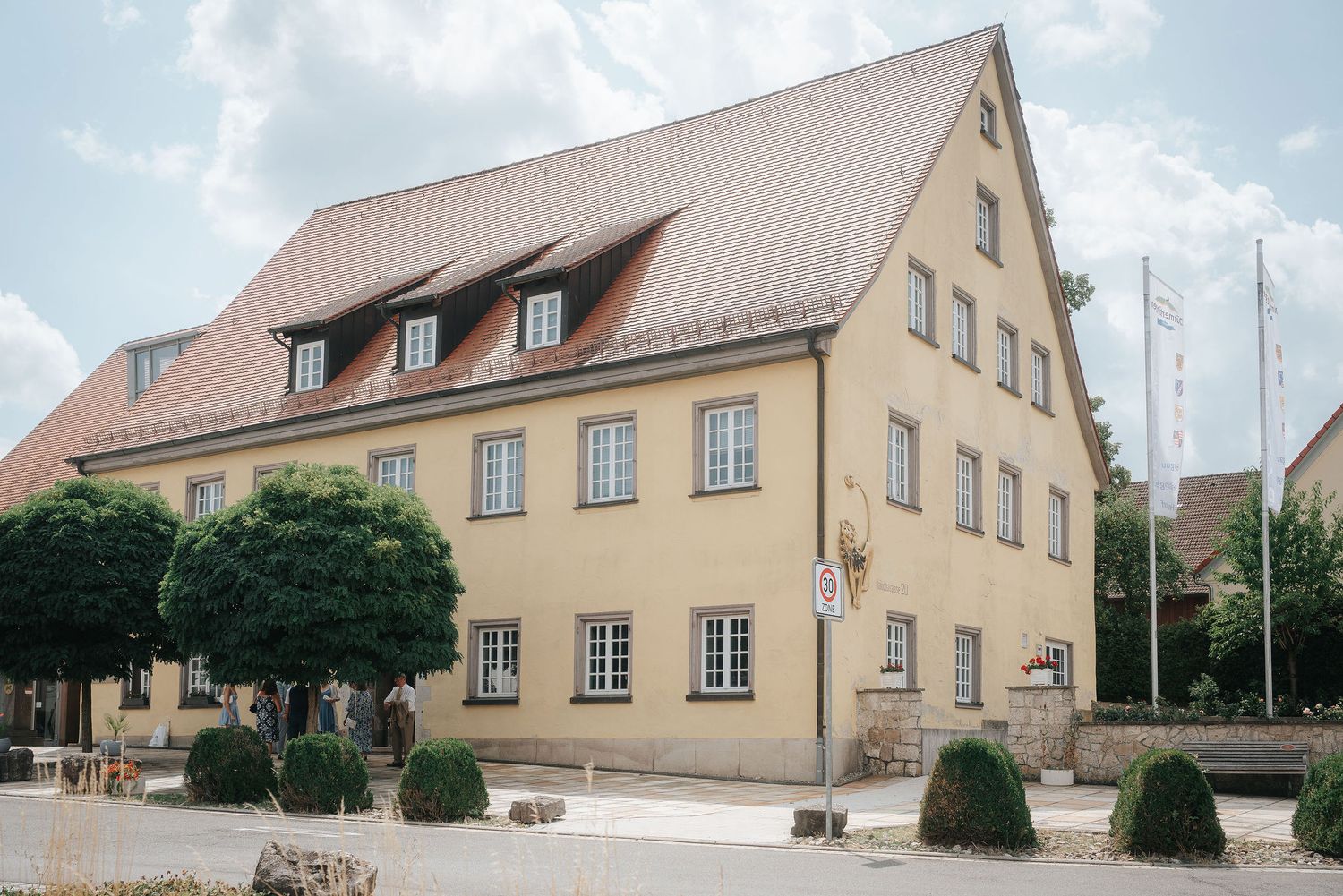 Historic yellow two-story building with a steep tiled roof and dormer windows, trimmed shrubs and trees in front, and flagpoles beside it.