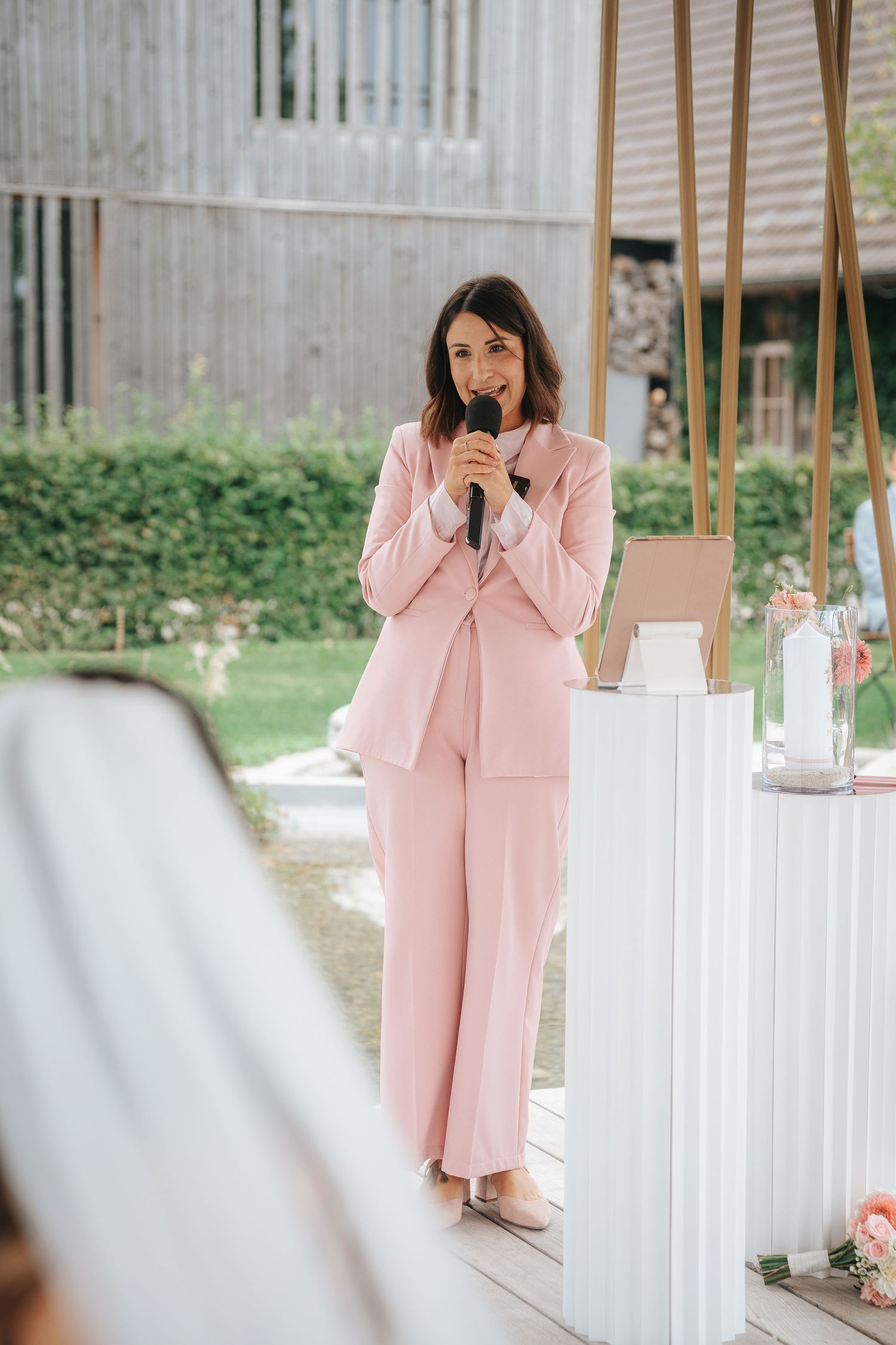 Woman in a pink pantsuit speaks into a microphone during an outdoor wedding ceremony beside a white pedestal with flowers.