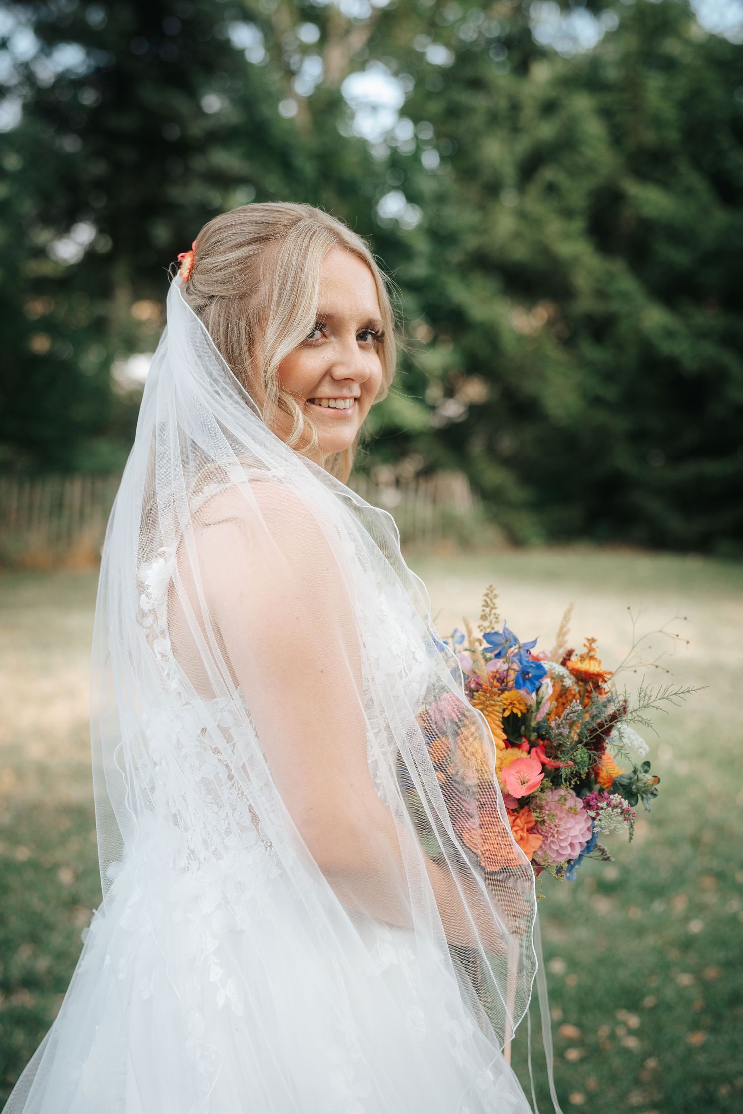 Smiling bride in a white lace wedding dress and long veil holds a colorful wildflower bouquet outdoors among trees.