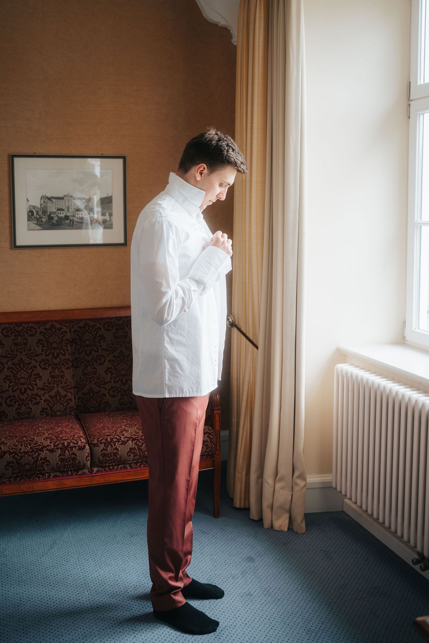 A man in a white shirt and burgundy trousers stands by a sunlit window, buttoning his collar in a classic hotel room with a red sofa.
