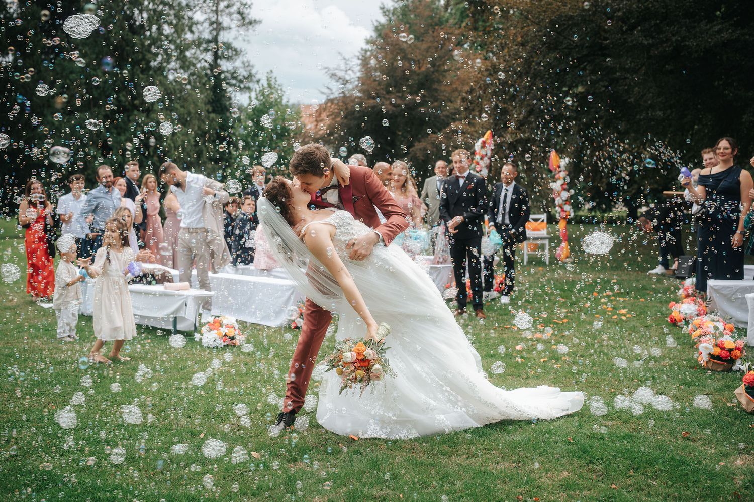 Bride and groom share a dramatic dipped kiss on a sunny lawn as wedding guests cheer among floating soap bubbles.