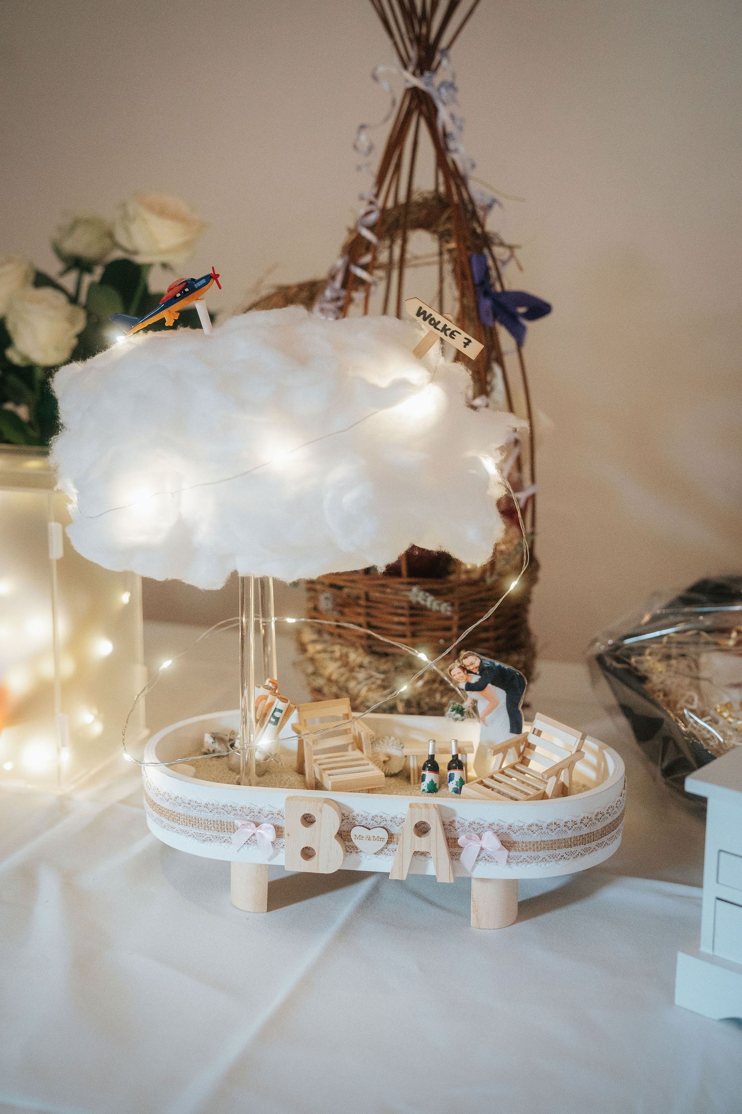 Handmade wedding centerpiece with a fluffy cloud, fairy lights, tiny bride and groom figurines and wooden BA letters on a round wooden base.