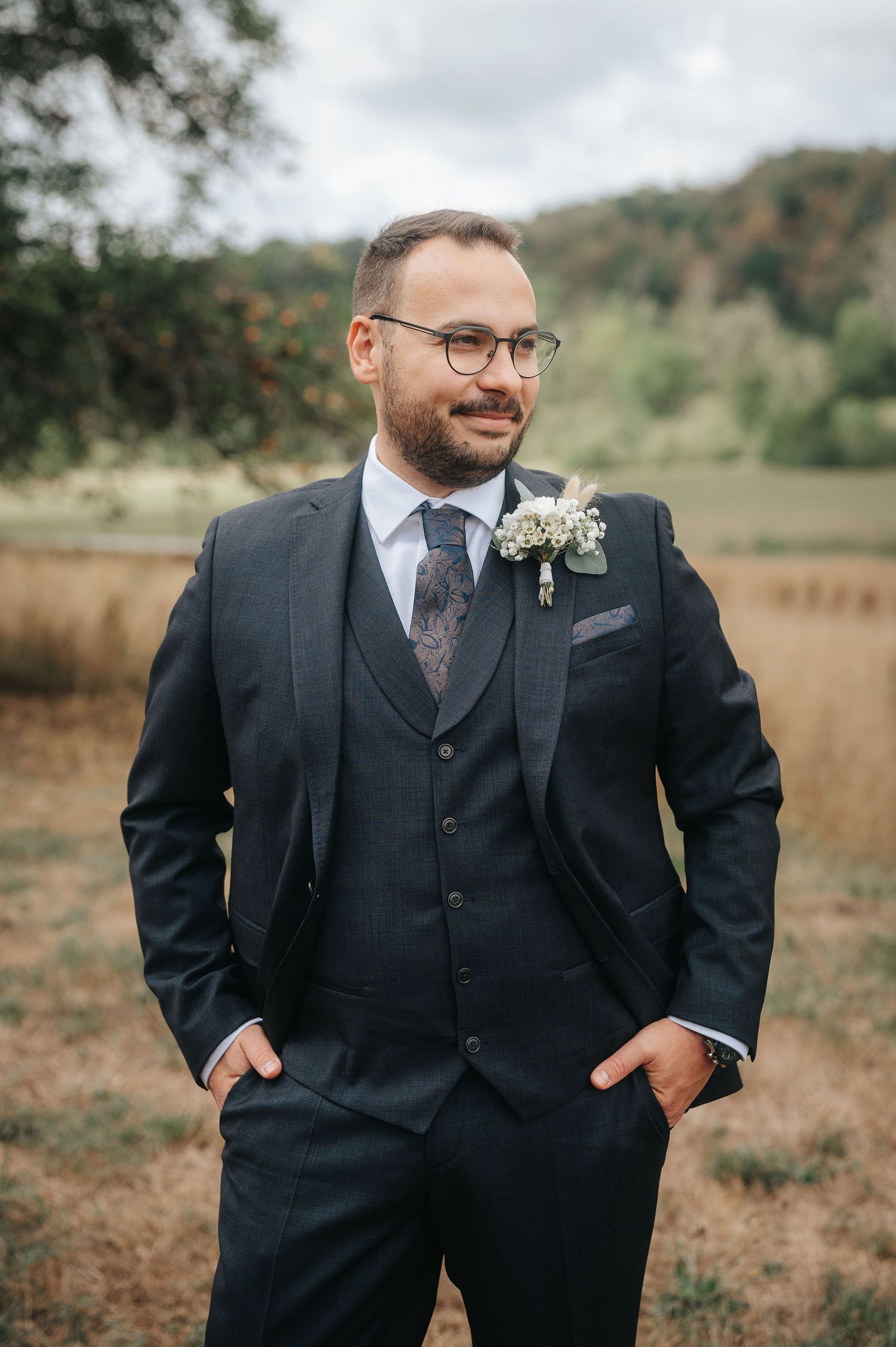 Smiling groom with glasses and a beard wears a dark three-piece suit with a floral boutonniere, standing with hands in pockets outdoors.