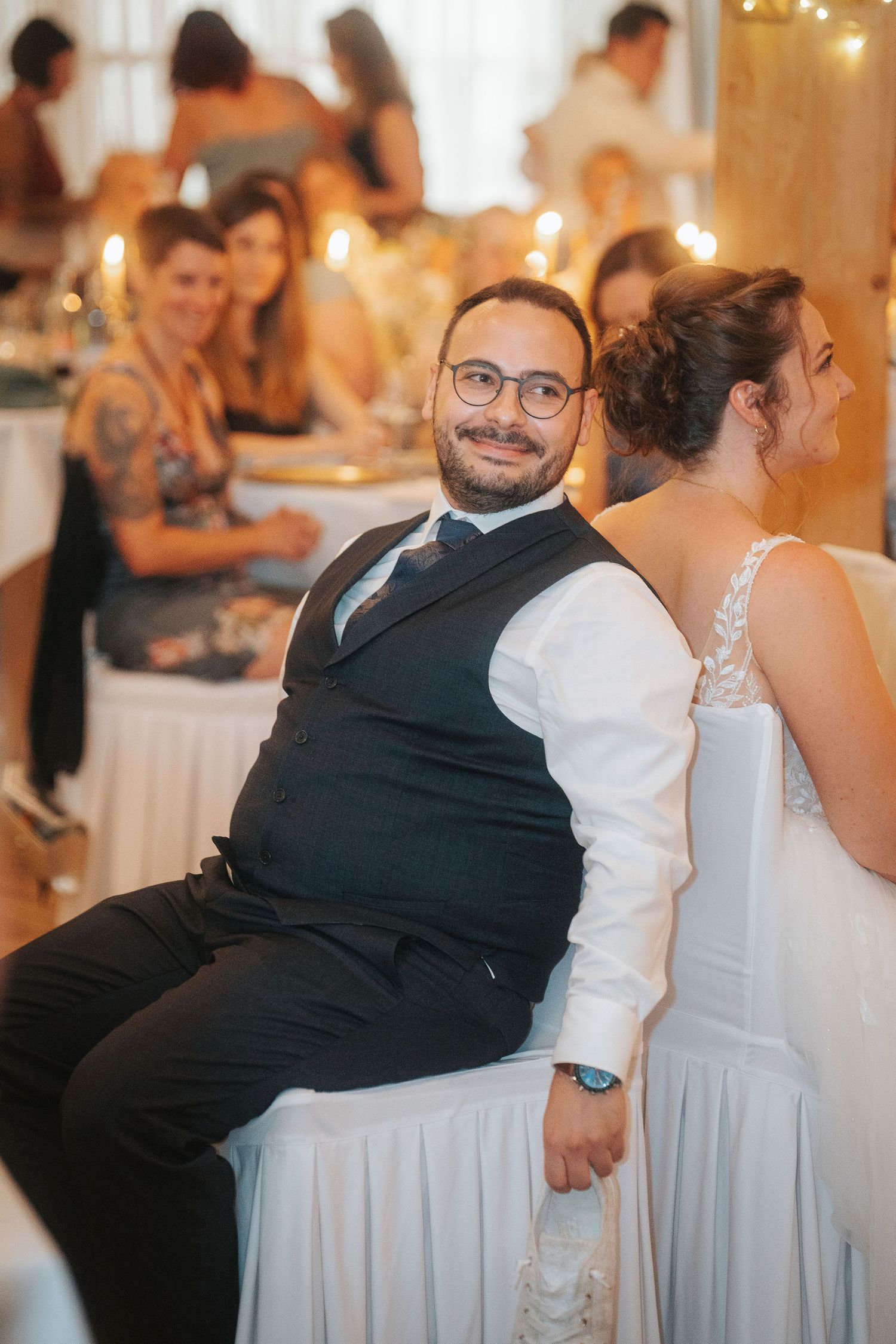 Smiling man in glasses, vest and tie sits beside a woman in a white dress at a candlelit wedding reception with guests behind.
