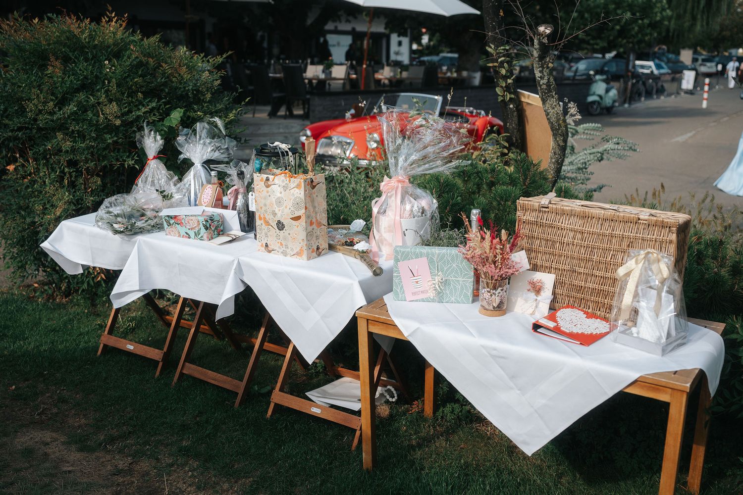Outdoor gift table with white tablecloths displaying wrapped treats, wicker baskets and handmade items on a grassy lawn near parked cars.