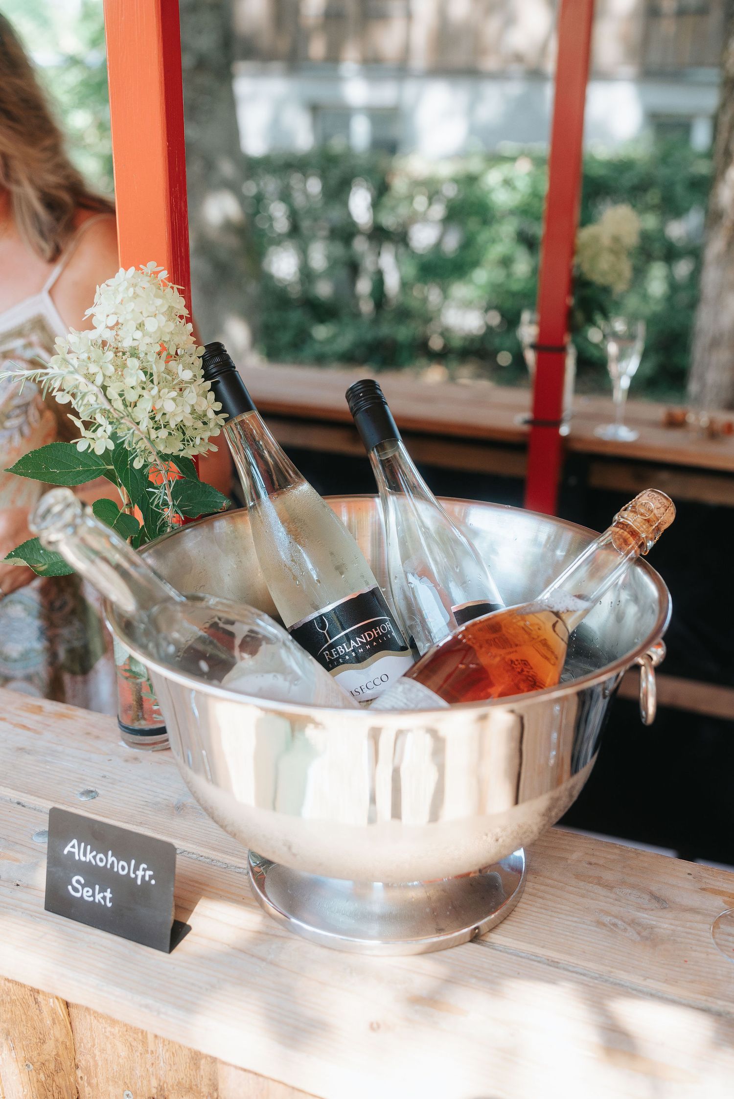 Silver ice bucket holding bottles of alcohol-free sparkling wine on a rustic wooden table beside white flowers and a chalkboard sign.