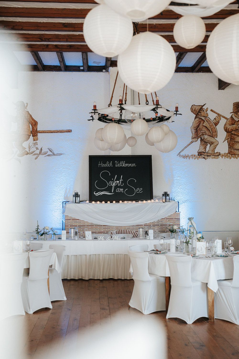 Wedding reception venue decorated with white paper lanterns, chair covers and chalkboard sign.