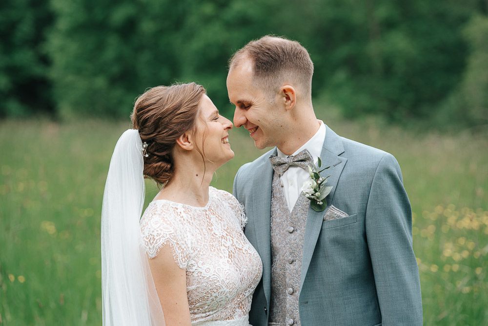 A wedding couple embraces in an outdoor meadow setting, with lush green grass and wildflowers surrounding them.
