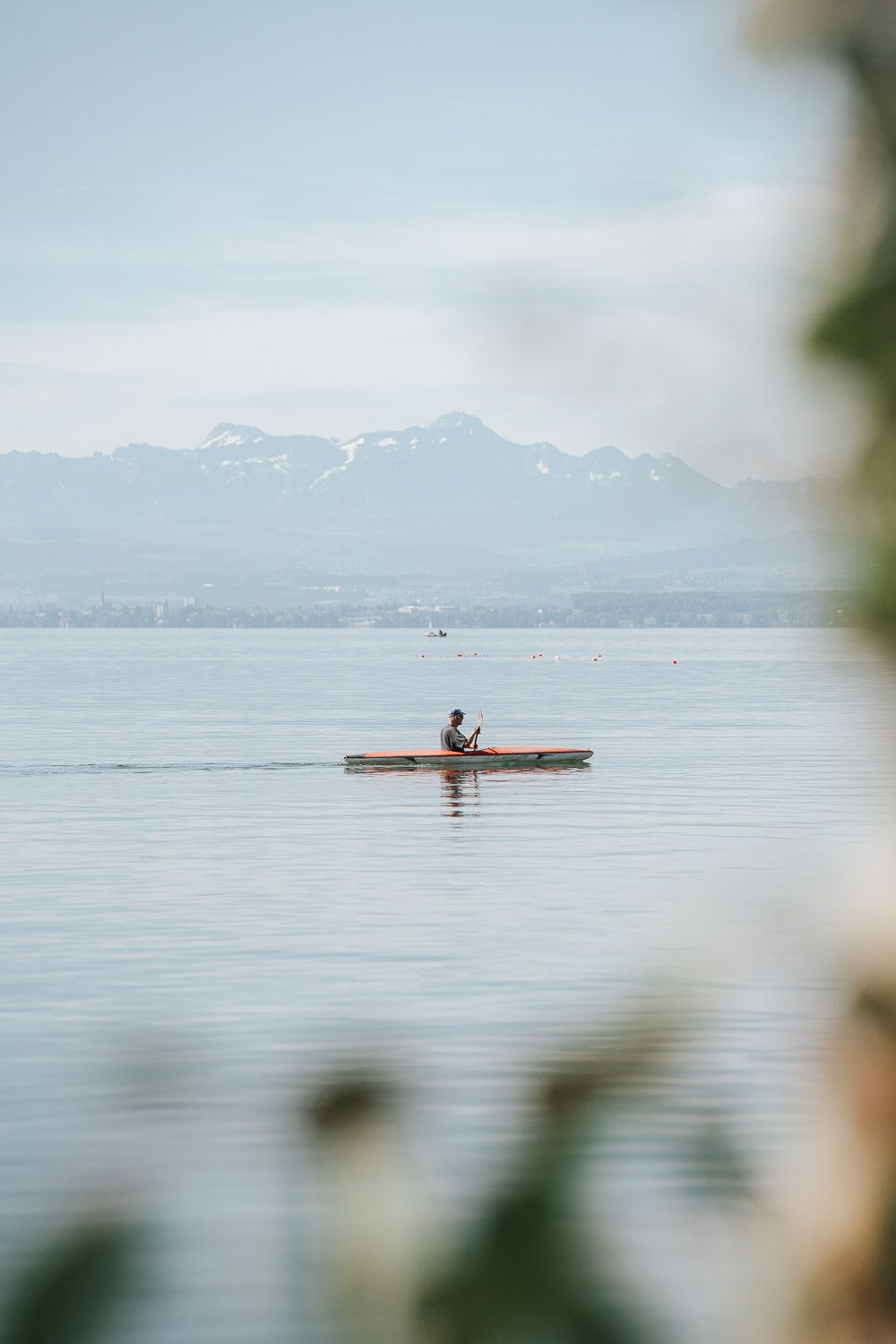 A kayaker paddles on calm water with snow-capped mountains in the distance, viewed through foliage in the foreground.