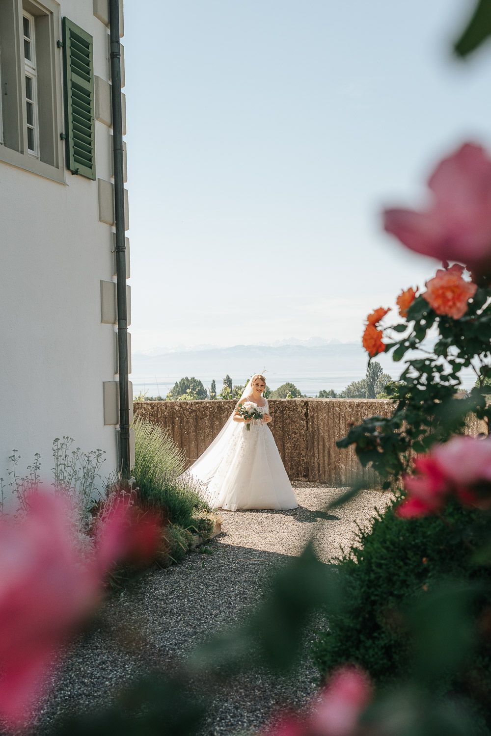 Bride in white wedding dress poses in garden with pink flowers and bamboo fence backdrop in sunlight.