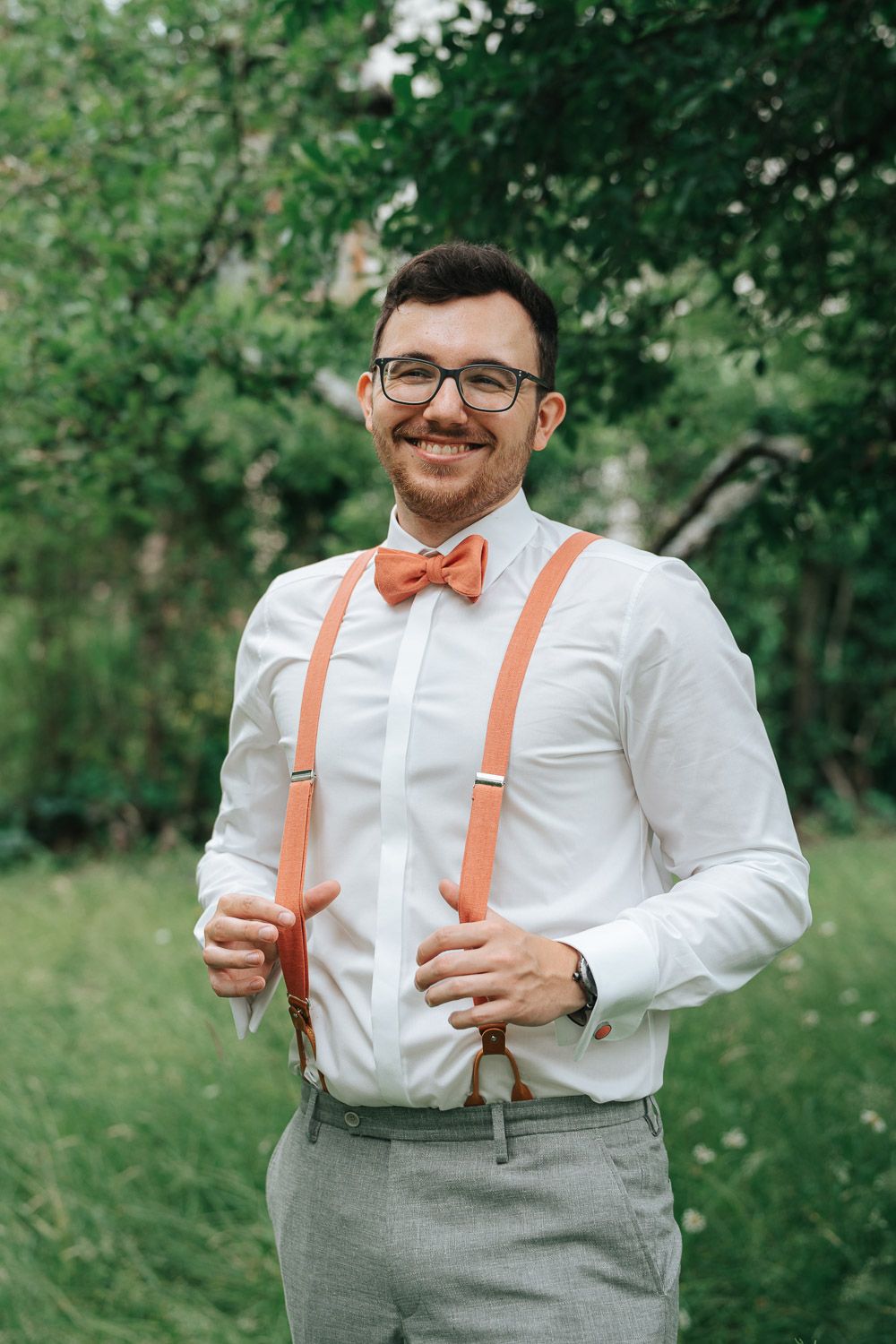 A person wearing a white dress shirt, gray slacks, orange suspenders and bow tie smiles outdoors in a garden setting.