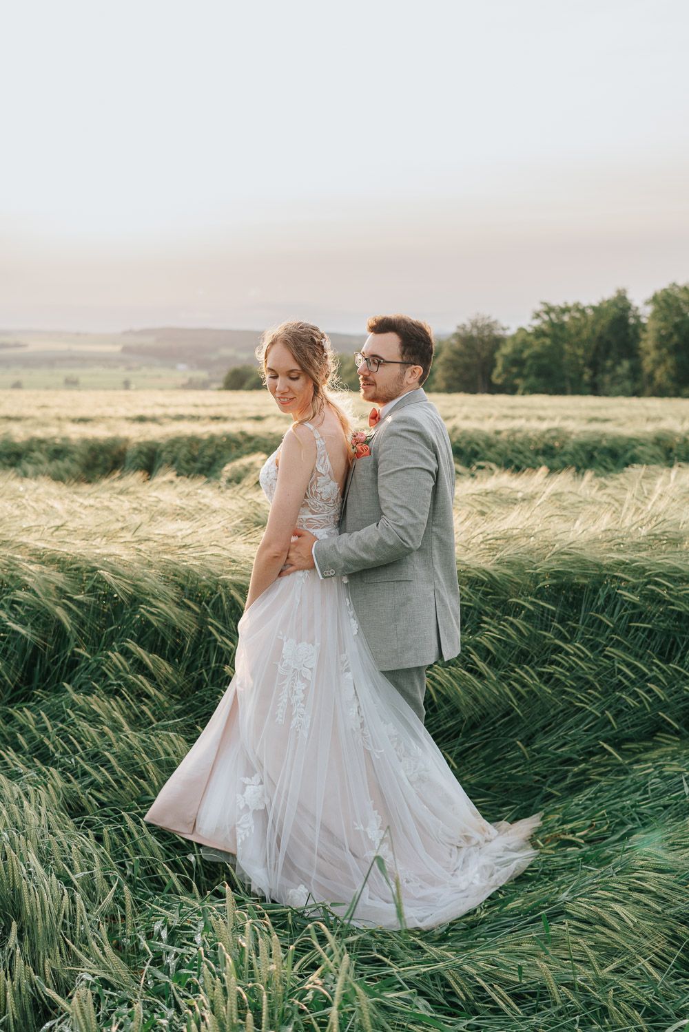 Wedding couple poses elegantly in a rural field with flowing tall grass and distant hills.