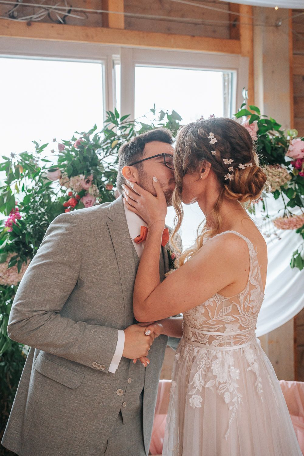 A romantic wedding moment captured as couple shares first kiss under floral decor in sunlit indoor venue.