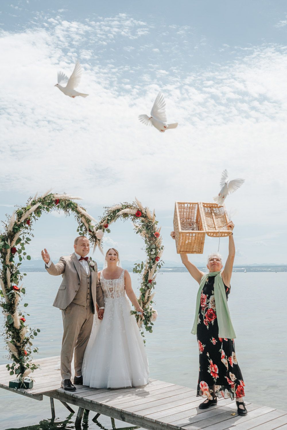 Wedding ceremony on wooden dock by water with flower arch and white doves being released from cage into blue sky.