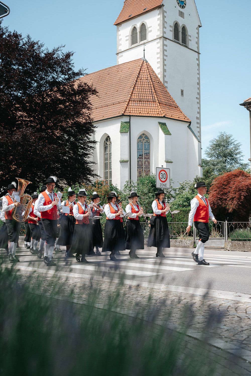 Traditional band members in Bavarian folk costumes perform outside a white church with a tall tower in the background.