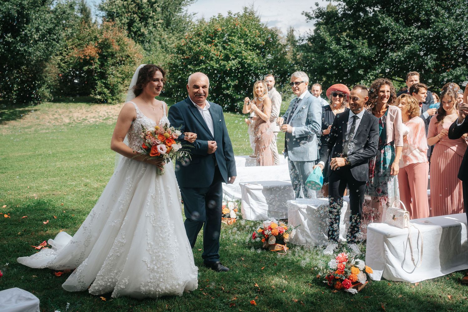 Bride in a white lace gown holding an orange bouquet walks down the aisle on the arm of an older man at an outdoor garden wedding ceremony.