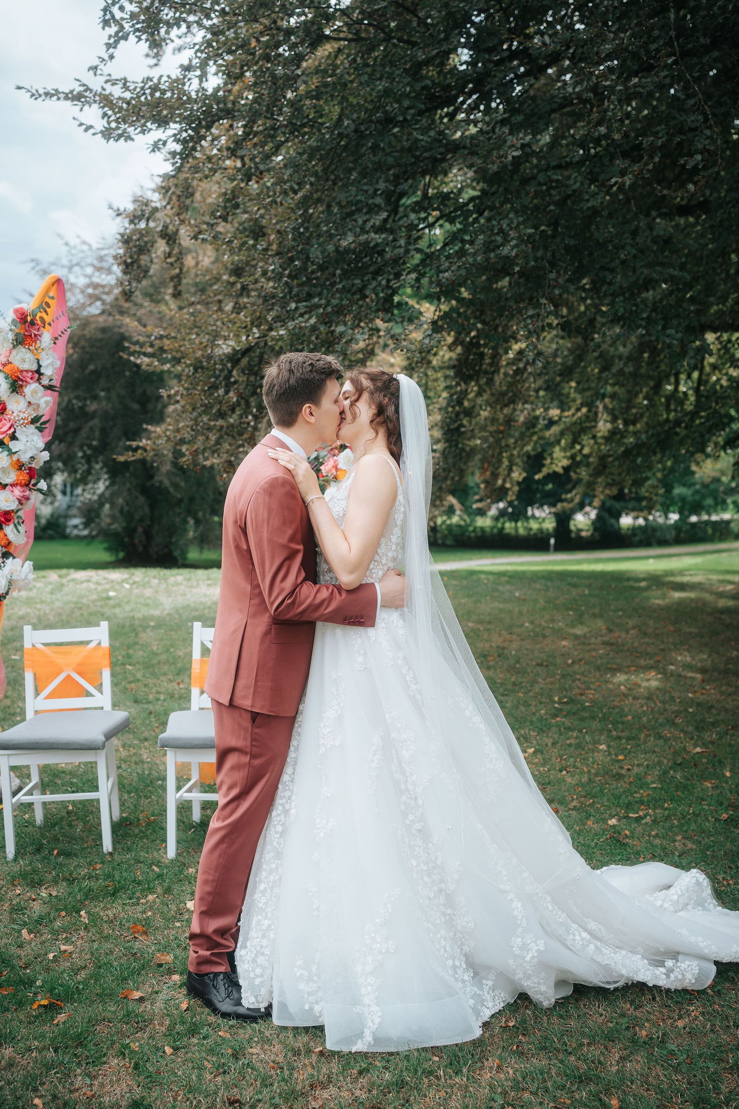 Bride in a lace wedding gown and veil kisses groom in a rust suit during an outdoor garden ceremony beside floral arch and white chairs.