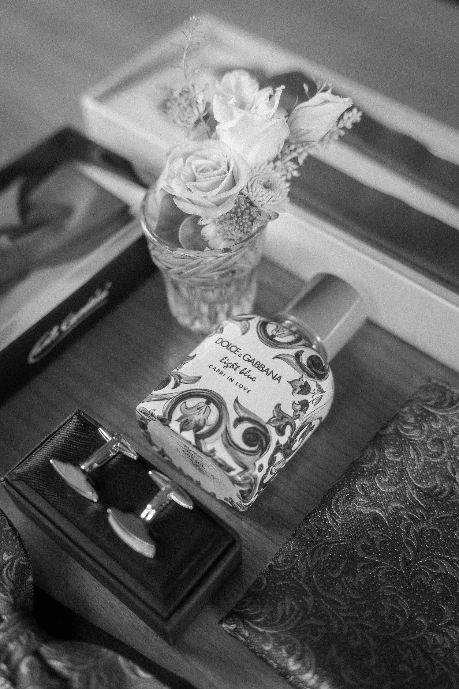 Black and white photo of a Dolce & Gabbana Light Blue perfume bottle beside cufflinks, a small rose bouquet and a brocade pocket square.