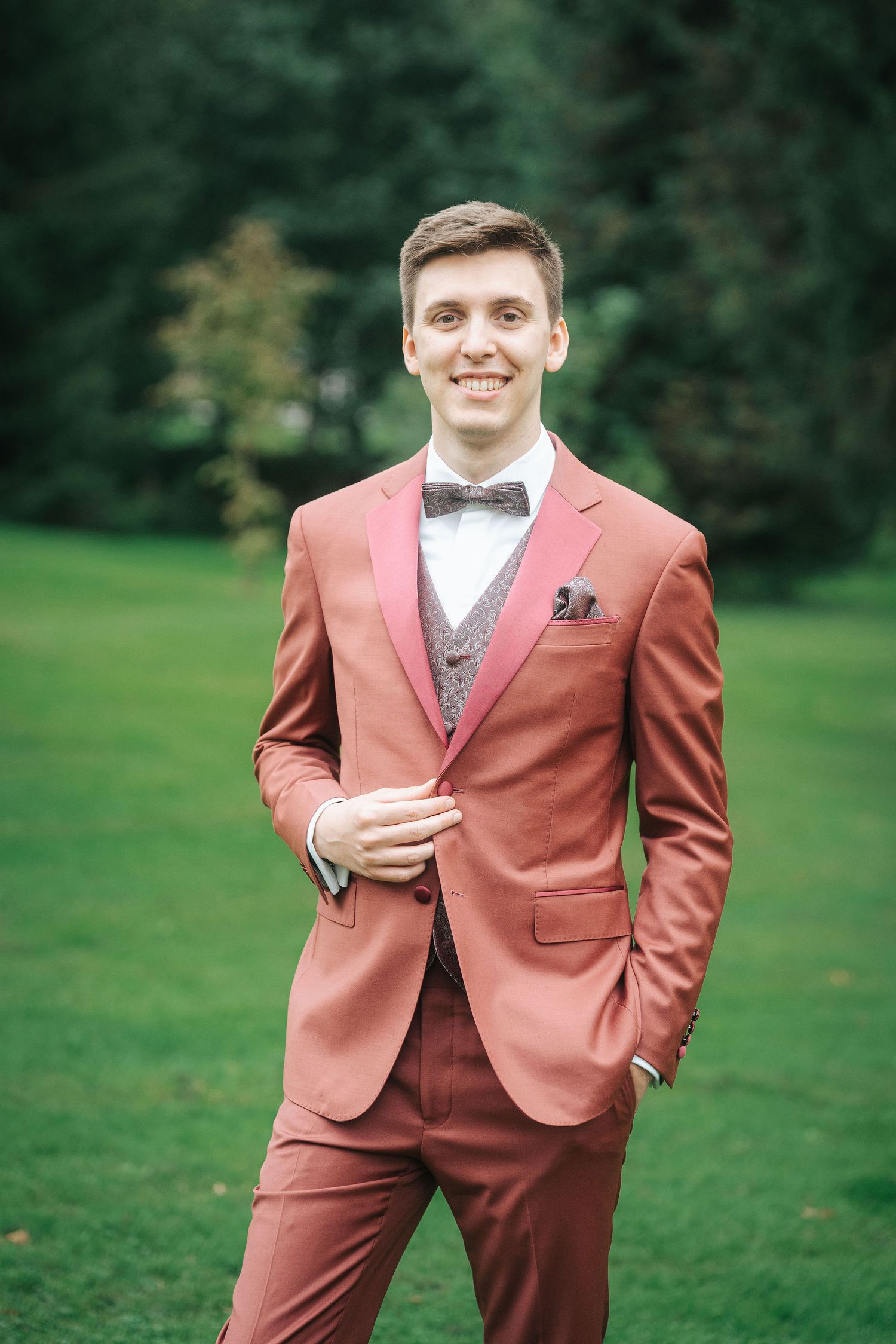 Smiling man in a rust-colored suit with bow tie, gray vest and pocket square stands on a green lawn with trees behind him.