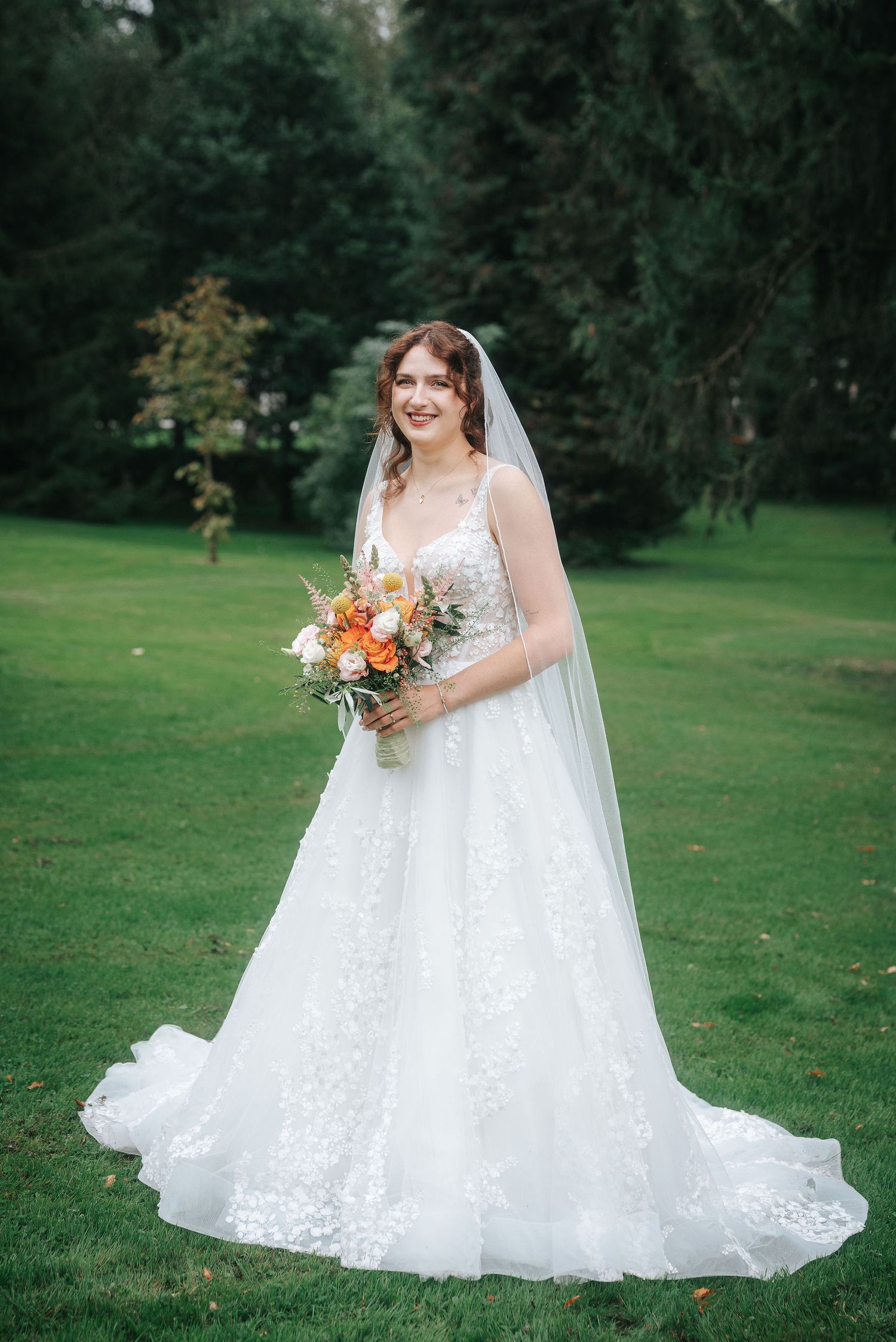 Smiling bride in a lace A-line wedding gown and long veil holds an orange and white floral bouquet on a green lawn surrounded by trees.